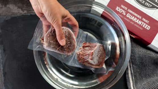 Person placing two sealed frozen Omaha Steaks filet mignon steaks in a bowl of cold water