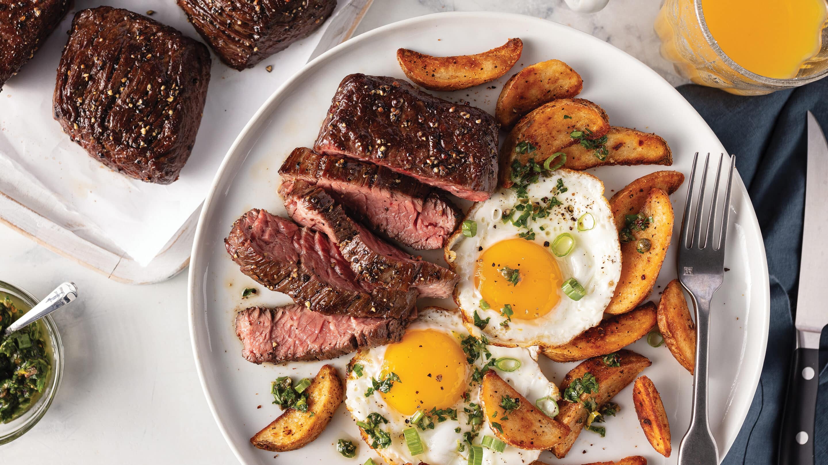 White plate with sliced steak, 2 over-easy eggs, potato wedges. Served with orange juice.