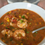 Red shrimp, corn, and tomato stew in white bowl with spoon