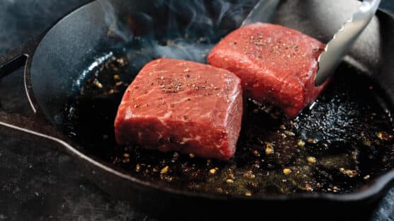 Two uncooked top sirloin steaks being placed in a hot cast iron skillet