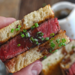 man holding wagyu filet mignon sandwich with green onion and pan sauce garnish