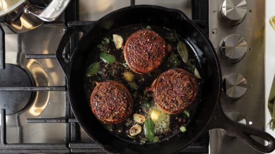 Three bacon-wrapped filet mignon steaks cooking in a cast iron skillet with garlic cloves, butter, and fresh herbs.