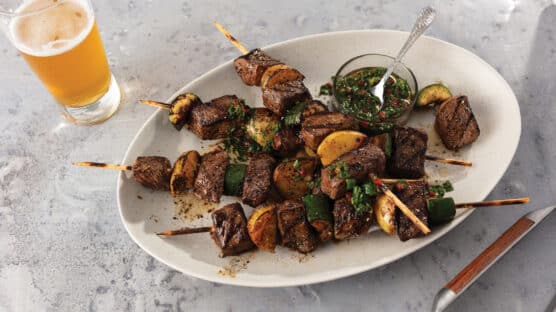 Steak and potato kabobs served on a white platter with a glass of light beer