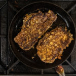 Two potato chip encrusted New York strip steaks in a cast iron skillet on stove
