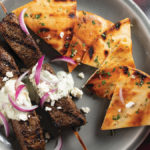 Two Mediterranean-style grilled Filet Mignon steak kabobs on bamboo skewers covered with tzatziki sauce and sliced red onions and served with grilled seasoned pita bread