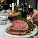 Dijon-Herb Prime Rib Roast sliced on platter and served with garlic butter mushrooms and red wine on a table set for a holiday gathering