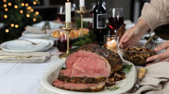 Dijon-Herb Prime Rib Roast sliced on platter and served with garlic butter mushrooms and red wine on a table set for a holiday gathering