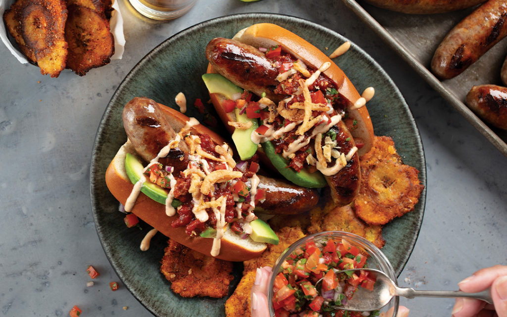 2 Mexican Bratwurst Dogs with grilled bratwurst in a toasted bun topped with chorizo chili, avocado slices, pico de gallo, chipotle aioli, and crispy fried shallots. Served on a silver platter with BBQ Tostones.