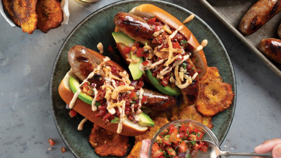 2 Mexican Bratwurst Dogs with grilled bratwurst in a toasted bun topped with chorizo chili, avocado slices, pico de gallo, chipotle aioli, and crispy fried shallots. Served on a silver platter with BBQ Tostones.