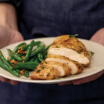 Woman holding white plate with sliced air fried chicken breast and green beans with bacon