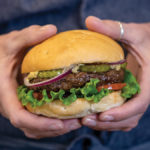 Person who holding hamburger with air fried burger, lettuce, tomato, pickles and red onion
