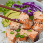 White bowl with white rice, cubed and air-fried bang bang salmon, avocado slices, pickled red onions, and green onions.