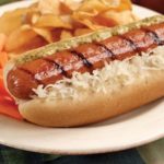 Omaha Dog with grilled gourmet frank, sauerkraut and Thousand Island dressing served with chips and carrots sticks on white plate