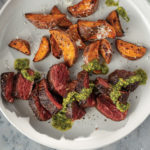 White plate with sliced bourbon brown sugar grilled hanger steak with drizzled pistachio pesto and grilled sweet potatoes. Served with side bowl of pistachio pesto.