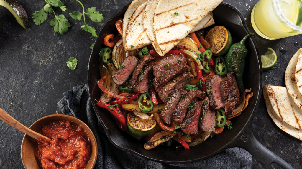 Cast iron pan with grilled bell peppers and lime wedges topped with sliced, grilled bavette steak cooked to a medium-rare doneness. Served with grilled tortillas and a cup of salsa.