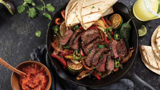 Cast iron pan with grilled bell peppers and lime wedges topped with sliced, grilled bavette steak cooked to a medium-rare doneness. Served with grilled tortillas and a cup of salsa.