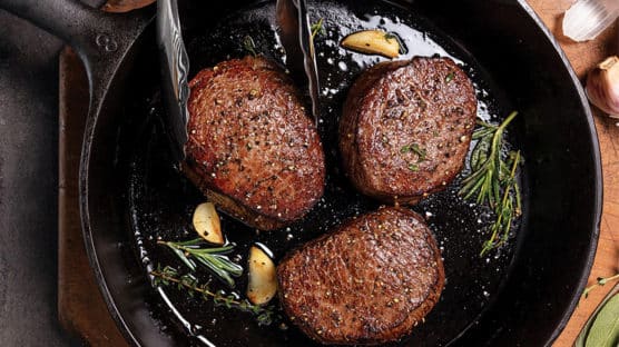 Three filet mignon steaks with great crust, garlic cloves, and fresh herbs cooking in a cast iron pan