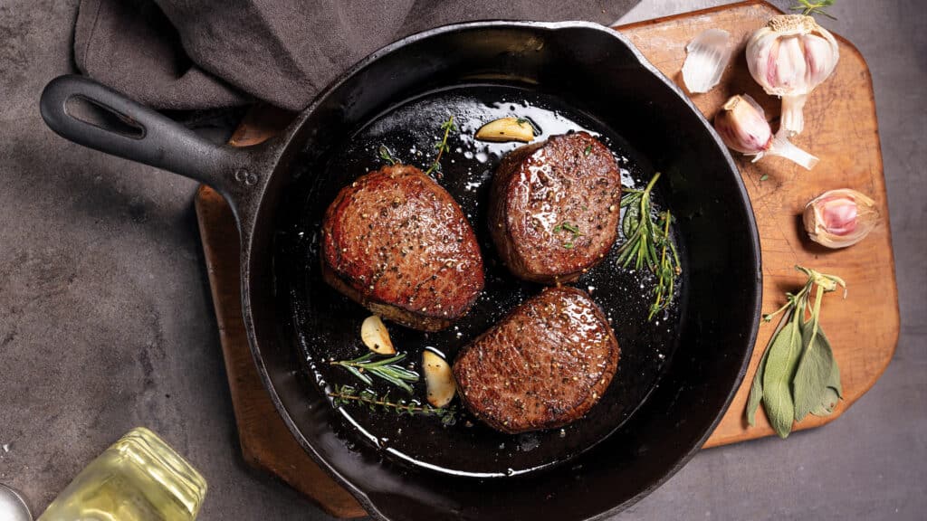 Cast iron skillet on cutting board with three pan-seared filet mignon with garlic cloves and fresh herbs.