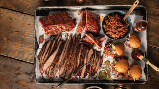 Large platter with smoked ribs, pulled pork in a bowl, pulled pork sandwiches, pickles and sliced brisket