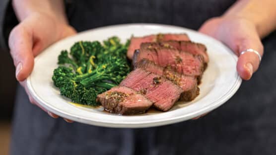 Chef holding white plate with cooked, sliced filet mignon served with a side of broccolini.