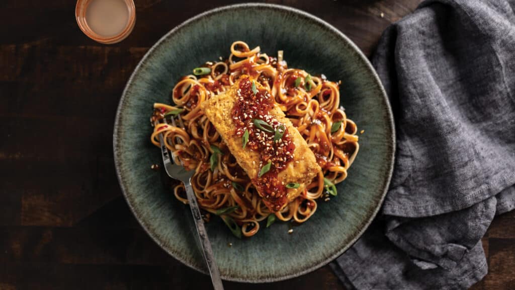 Grey plate Sweet 'n sour pan-roasted halibut with sauce on a bed of sesame butter noodles next to a blue napkin and drink glass.