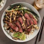 Korean-style steak bowl with coconut rice, spicy Korean mayo, sesame mushrooms and marinated ribeye crown steak in white bowl on table.