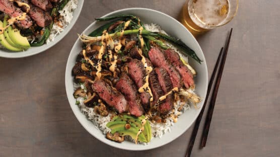 Korean-style steak bowl with coconut rice, spicy Korean mayo, sesame mushrooms and marinated ribeye crown steak in white bowl on table.