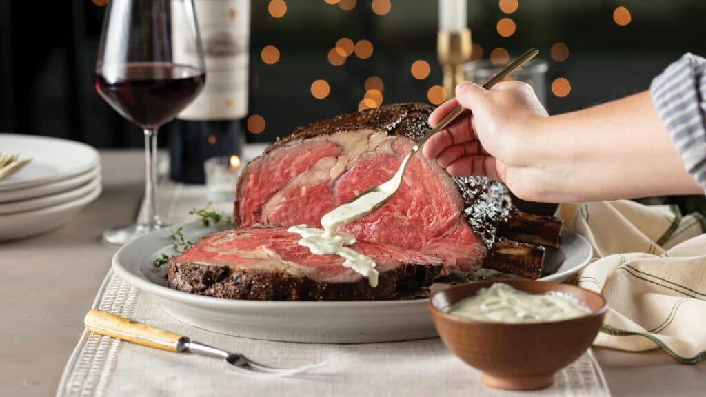 Holiday table with medium-rare prime rib roast sliced with person adding a dollop of jalapeno crema.