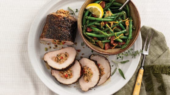 Sausage-stuffed pork roast sliced and plated with a bowl of blistered green beans with bacon