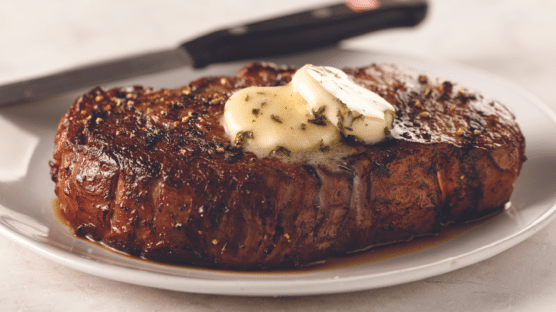 Cooked ribeye steak with 2 pats of compound butter on white plate with steak knife in background.