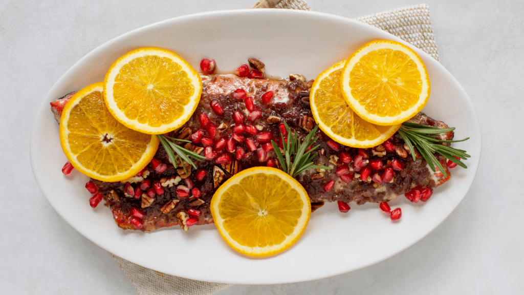 Side of cooked salmon plated with pomegranates, rosemary sprigs and sliced oranges.