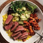 Mediterranean steak bowl salad with mixed green salad, turmeric rice, roasted rosemary sweet potatoes, and quick pickes topped with a medium-rare sliced bavette steak.