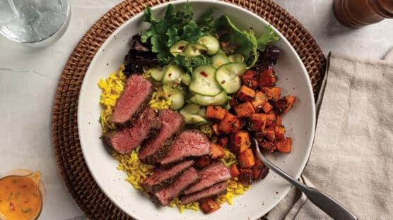 Mediterranean steak bowl salad with mixed green salad, turmeric rice, roasted rosemary sweet potatoes, and quick pickes topped with a medium-rare sliced bavette steak.