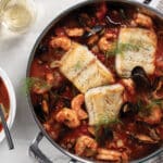 Stew pot and white white bowl on table filled with Sablefish Cioppino. Served with glass of white wine and garlic-parmesan bread.