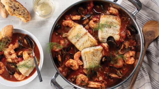 Stew pot and white white bowl on table filled with Sablefish Cioppino. Served with glass of white wine and garlic-parmesan bread.