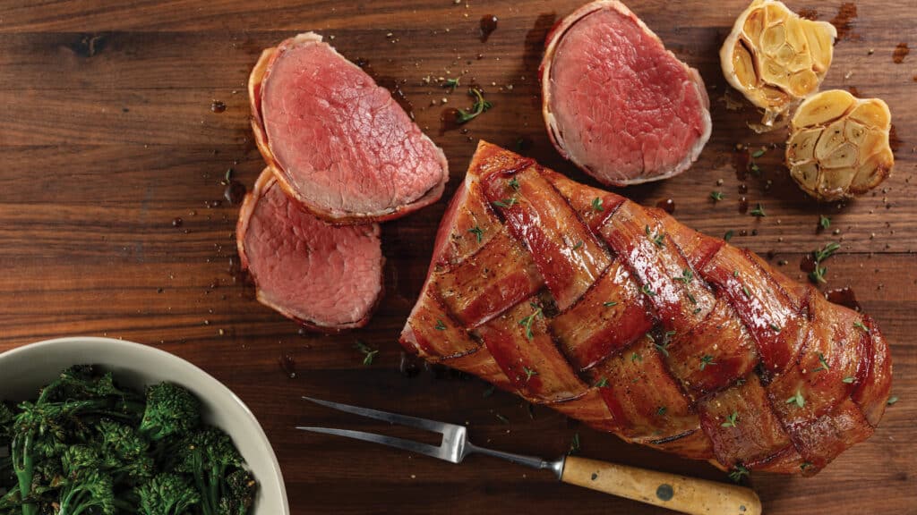 Cutting board with cooked bacon-wrapped chateaubriand roast with roasted garlic head and bowl of broccolini