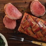 Cutting board with cooked bacon-wrapped chateaubriand roast with roasted garlic head and bowl of broccolini