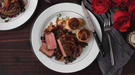 White plate with sliced pan-seared filet mignon, melted potatoes and stir-fried red chard on a romantic table setting with red roses.
