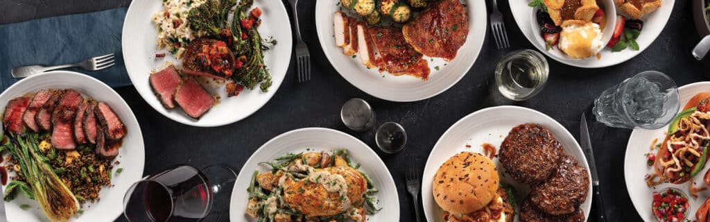 Table set with 7 different plates of Omaha Steaks food., including steak dinners, barbecue chicken, caramel apple tartlets, hot dogs, burgers, and chicken.