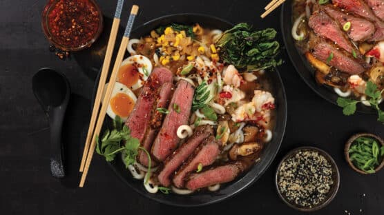 Udon noodle soup with sliced ribeye, lobster, soft boiled egg, and udon noodles in a black bowl with chopsticks.