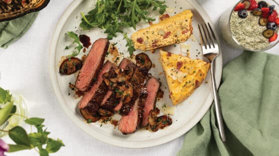 White plate with sliced Italian bavette steak, cooked medium-rare, and topped with balsamic mushrooms and served with potato-bacon fritatta and arugula.