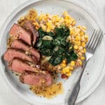 White plate on table with sliced medium-rare filet mignon on a bed of creamed corn and spinach, topped with a savory anchovy butter sauce