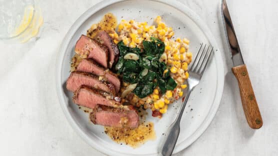 White plate on table with sliced medium-rare filet mignon on a bed of creamed corn and spinach, topped with a savory anchovy butter sauce