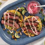 Two Grilled Flat Iron Steak Tostadas with Coconut-Chili Mayo and Guacamole served with pickled onions on a blue plate