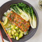 Teriyaki salmon bowl featuring Omaha Steaks salmon fillets, rice, bok choy, diced avocado, lime wedges in a grey bowl with chopsticks.