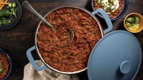 Table with crock of ground beef chili next to a bowl of chili garnished with jalapenos, diced green onions, and sour cream. Also on table is a bowl of sliced limes and a plate with sliced jalapenos.