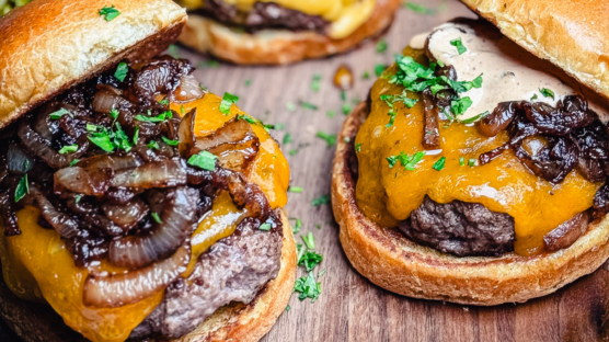 Two butter burgers topped with American cheese, caramelized onions, and garlic aioli on a rustic wood counter.