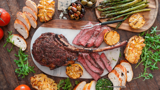 Board with tomahawk steak cooked medium rare, sliced bread, stuffed backed potatoes, arugula, stuffed olives, tomatotes, blue cheese, a bowl of chimichurri sauce, and charred lemons