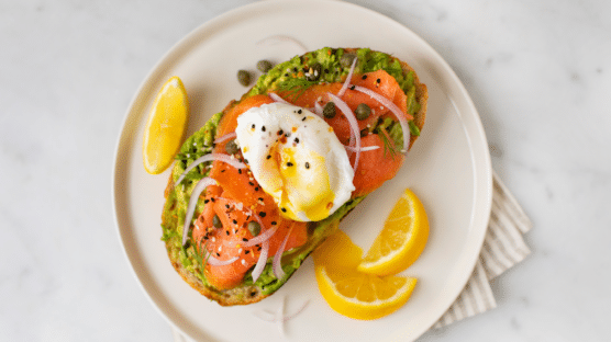 Avocado toast with cold-smoked salmon, sliced onion, and a poached egg on a white plate with orange slices.