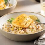 Table set with a white bowl with crab and corn chowder and topped with a pan-seared Chilean Sea Bass. Glass of Chardonnay and another bowl in the background.